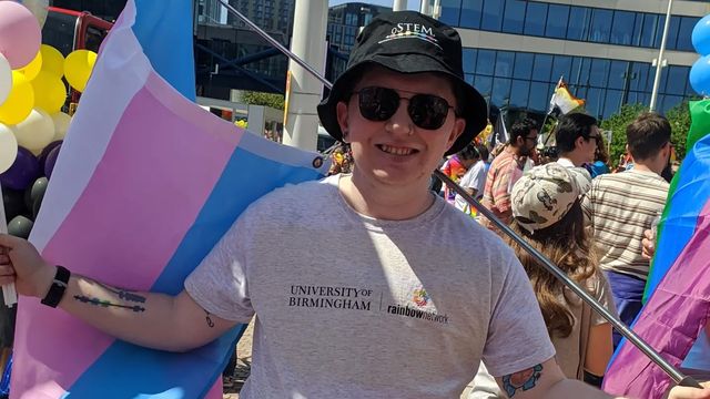 Avery Cunningham stands wearing a bucket hat and sunglasses and holding a transgender pride flag. 