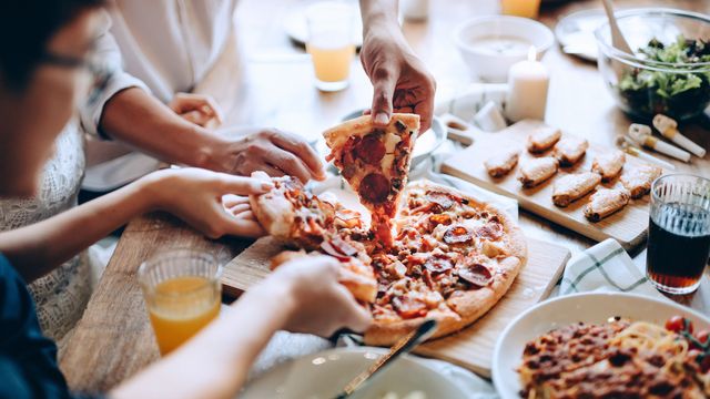 Three sets of hands pick up slices of pizza while sat at a table filled with other food items. 